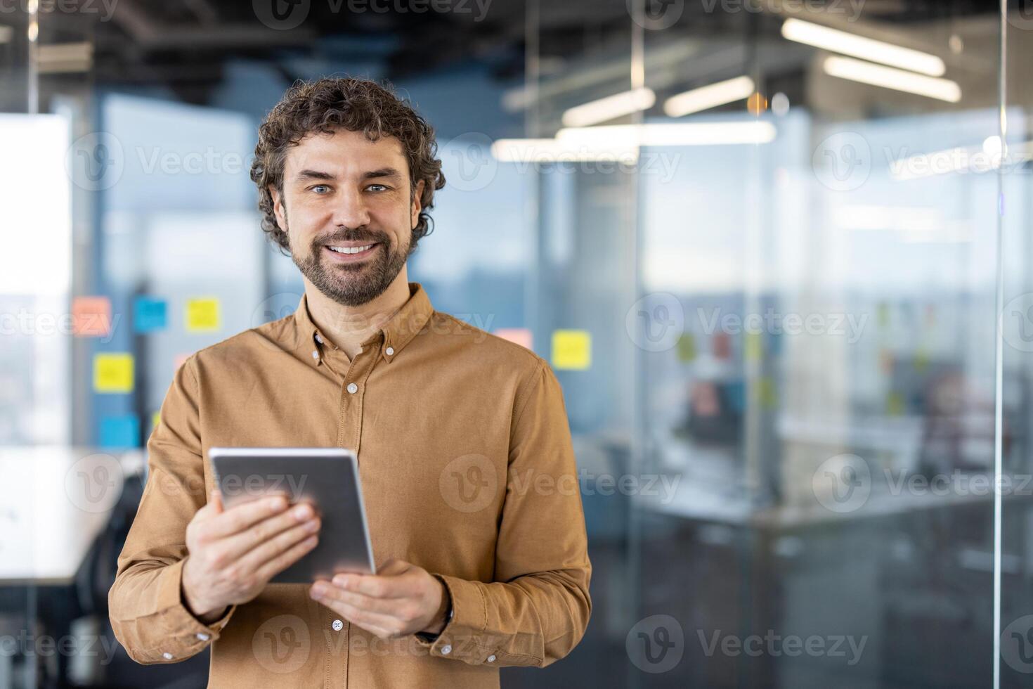 ritratto di un' maturo ispanico uomo d'affari sorridente con fiducia mentre Tenere un' digitale tavoletta nel un ufficio ambientazione con bicchiere muri. foto