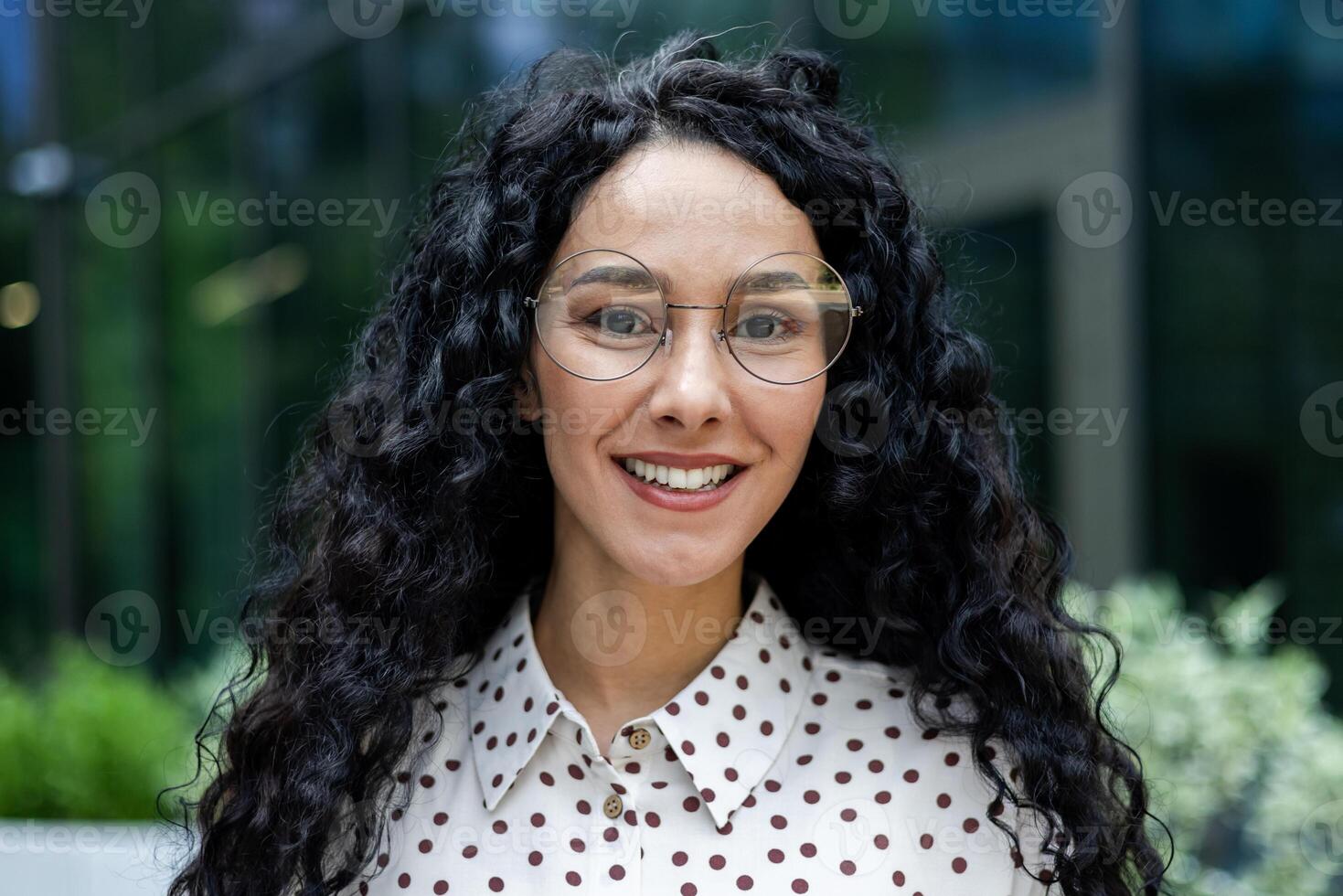 fiducioso professionale donna sta all'aperto, sua Riccio capelli fluente, indossare un' elegante polka punto camicetta, simboleggiante aziendale moda. foto