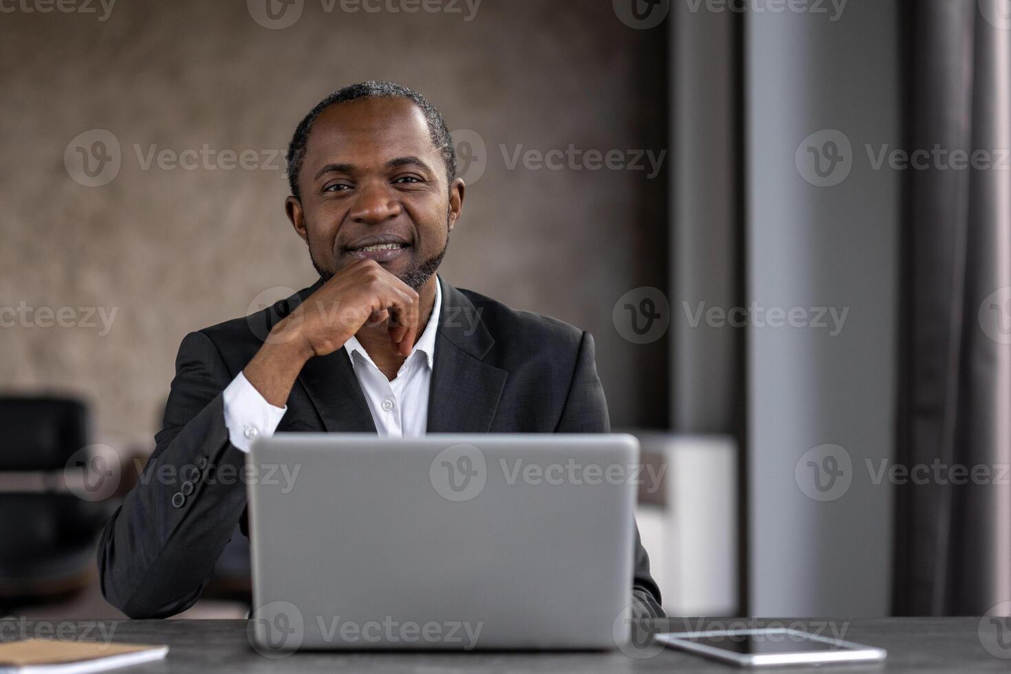un' professionale nero uomo d'affari sorrisi con fiducia nel un' moderno ufficio ambientazione, utilizzando un' il computer portatile a il suo scrivania circondato di tecnologia. foto