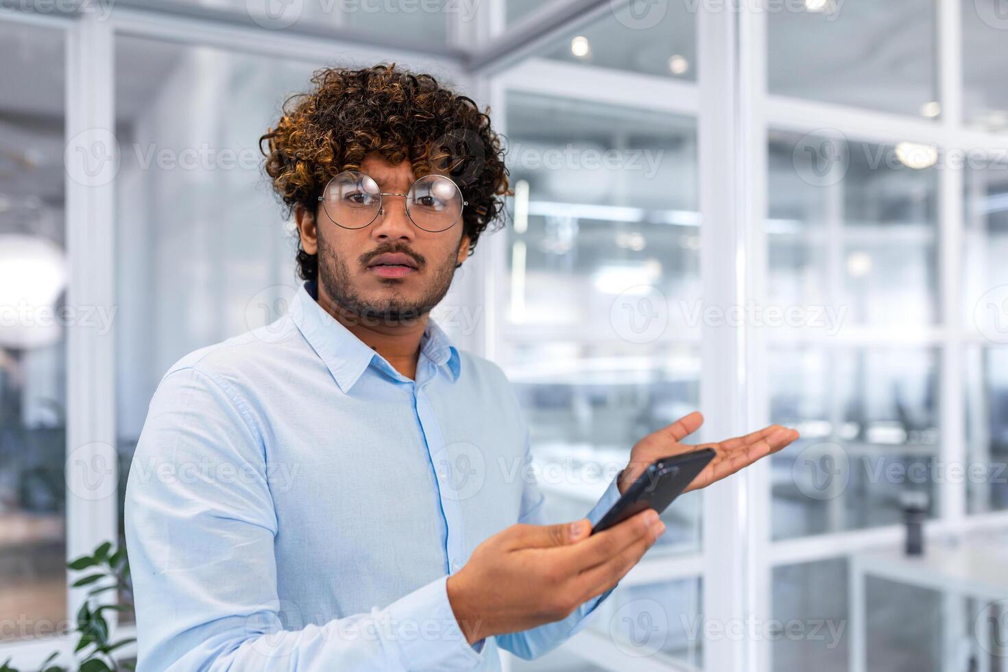 nervoso e insoddisfatto uomo con Telefono guardare a telecamera, giovane uomo d'affari indù programmatore Tenere smartphone, avuto risposta cattivo notizia e insoddisfacente opera risultati dentro ufficio. foto