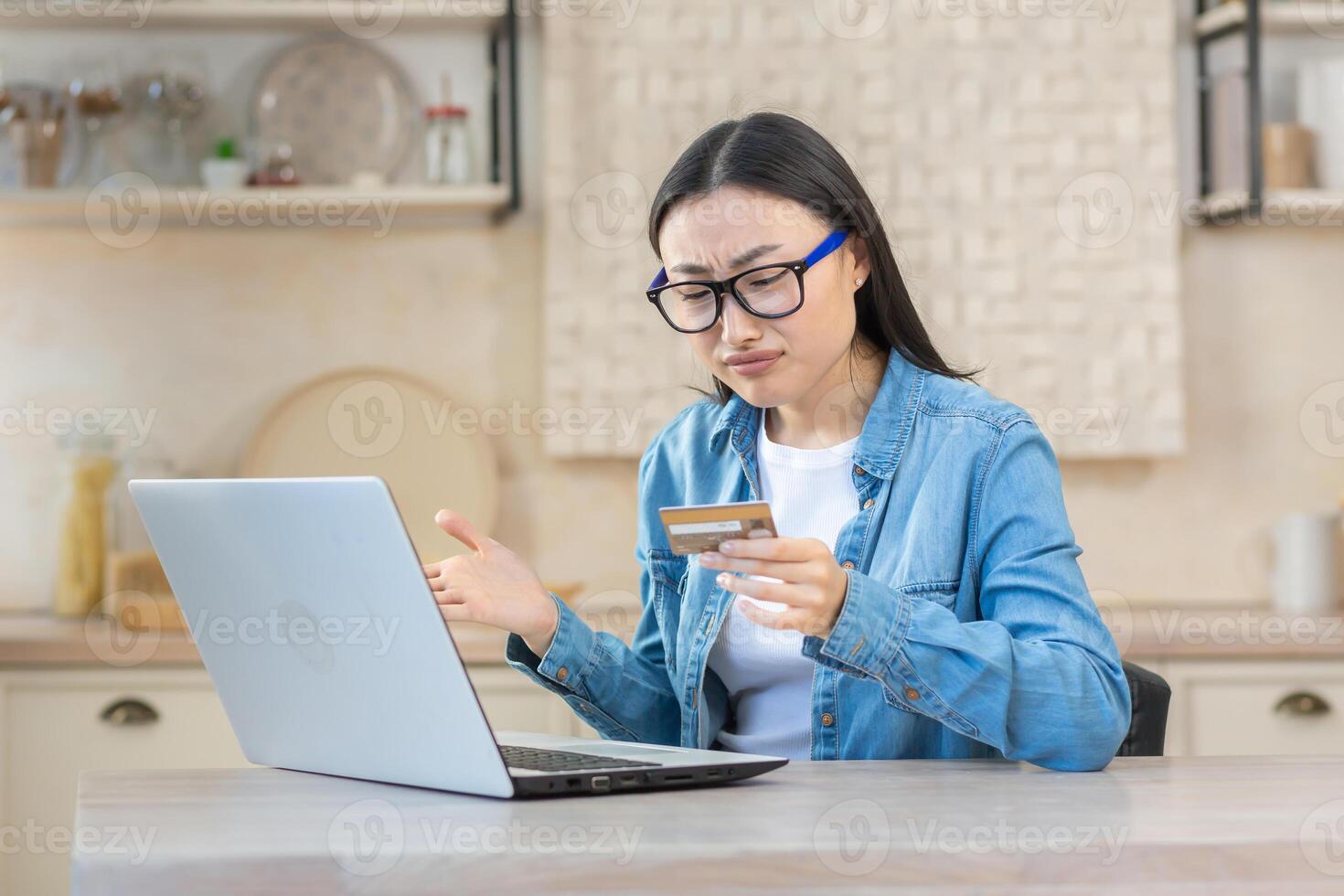 irritato e triste donna nel depressione provando per rendere in linea Acquista nel in linea negozio, asiatico donna seduta a casa nel cucina utilizzando il computer portatile e banca credito carta, incapace per rendere pagamento in linea. foto