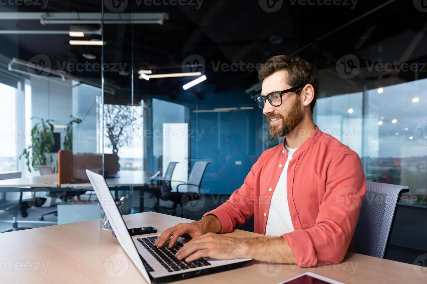 riuscito sorridente uomo Lavorando dentro ufficio con computer portatile, uomo d'affari nel rosso camicia sorridente e digitando su tastiera nel occhiali, programmatore Lavorando Software per programma. foto