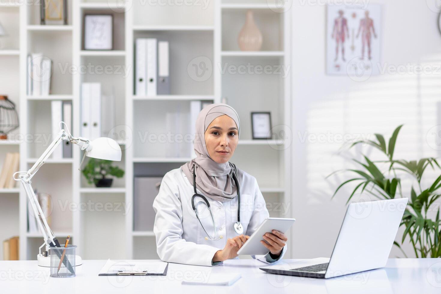 professionale musulmano femmina medico nel un' hijab e bianca cappotto Lavorando a sua ufficio, utilizzando un' tavoletta, con il computer portatile e stetoscopio su scrivania. foto
