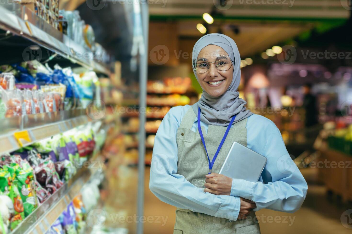 un' giovane donna, un' supermercato lavoratore nel un' hijab. lui sta vicino il contatori con merce, sorrisi foto