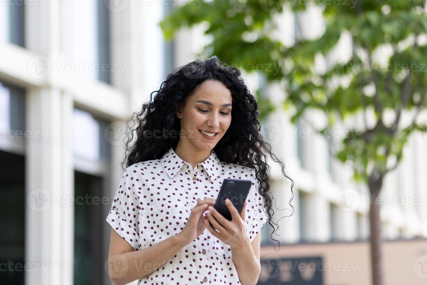 un' giovane bellissimo donna passeggiate attraverso il città con un' Telefono nel sua mani, sorrisi con soddisfazione, usi un' smartphone applicazione, sfoglia Internet pagine, tipi un' testo Messaggio, sociale reti. foto