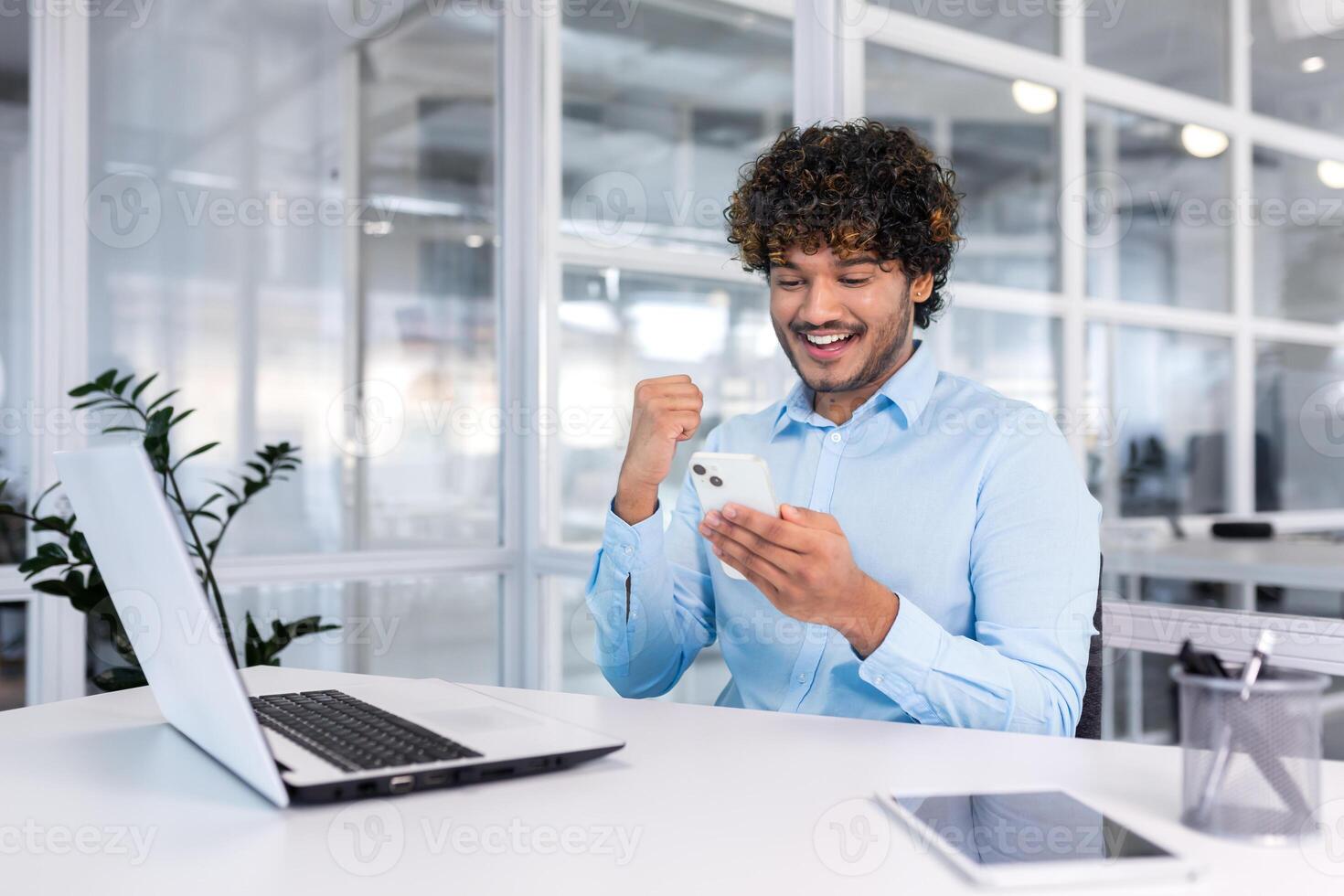 giovane ispanico uomo d'affari seduta nel il ufficio a il scrivania e utilizzando il Telefono. lui sembra a il schermo, quadranti, gioisce, festeggia, Spettacoli un' vittoria gesto con il suo mano. foto