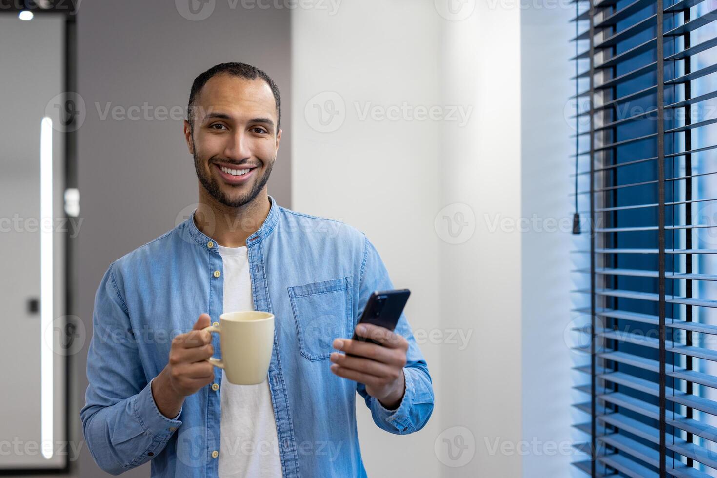 ritratto di un' sorridente musulmano uomo, un ufficio lavoratore e un' libero professionista, in piedi nel il sala di il finestra, Tenere un' tazza di caffè e un' mobile Telefono, guardare a il telecamera. foto