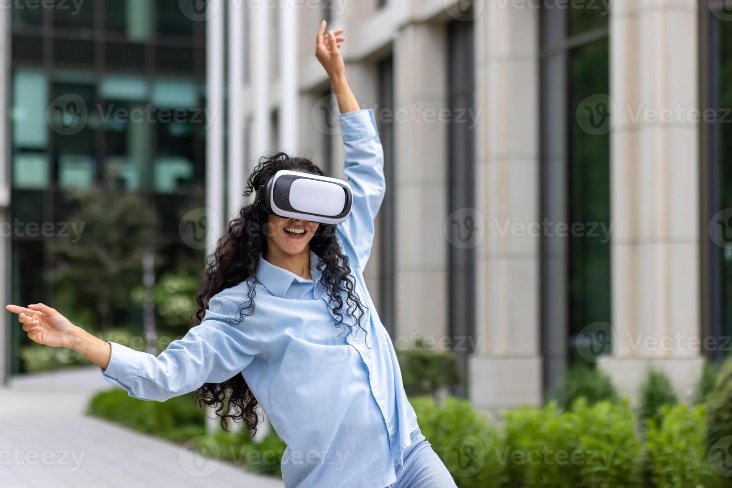 giovane bellissimo ragazza nel camicia e jeans danza al di fuori ufficio edificio indossare virtuale la realtà occhiali, ispanico donna contento avendo divertimento e rilassante con Riccio capelli. foto