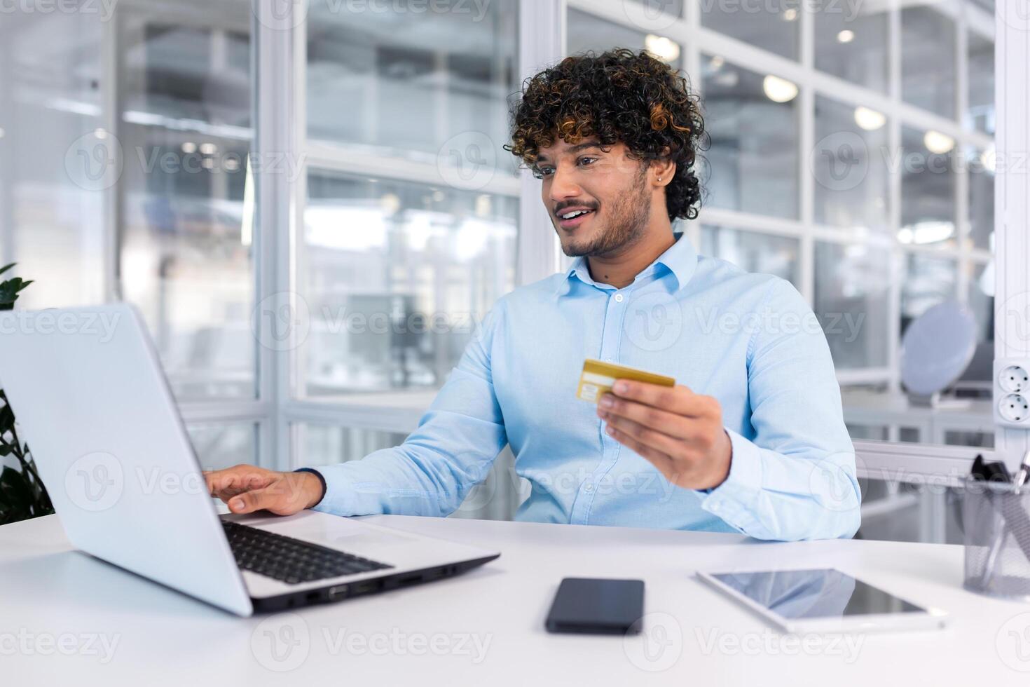 un' giovane ispanico uomo è seduta a un' scrivania nel un ufficio utilizzando un' il computer portatile e utilizzando un' credito carta. foto