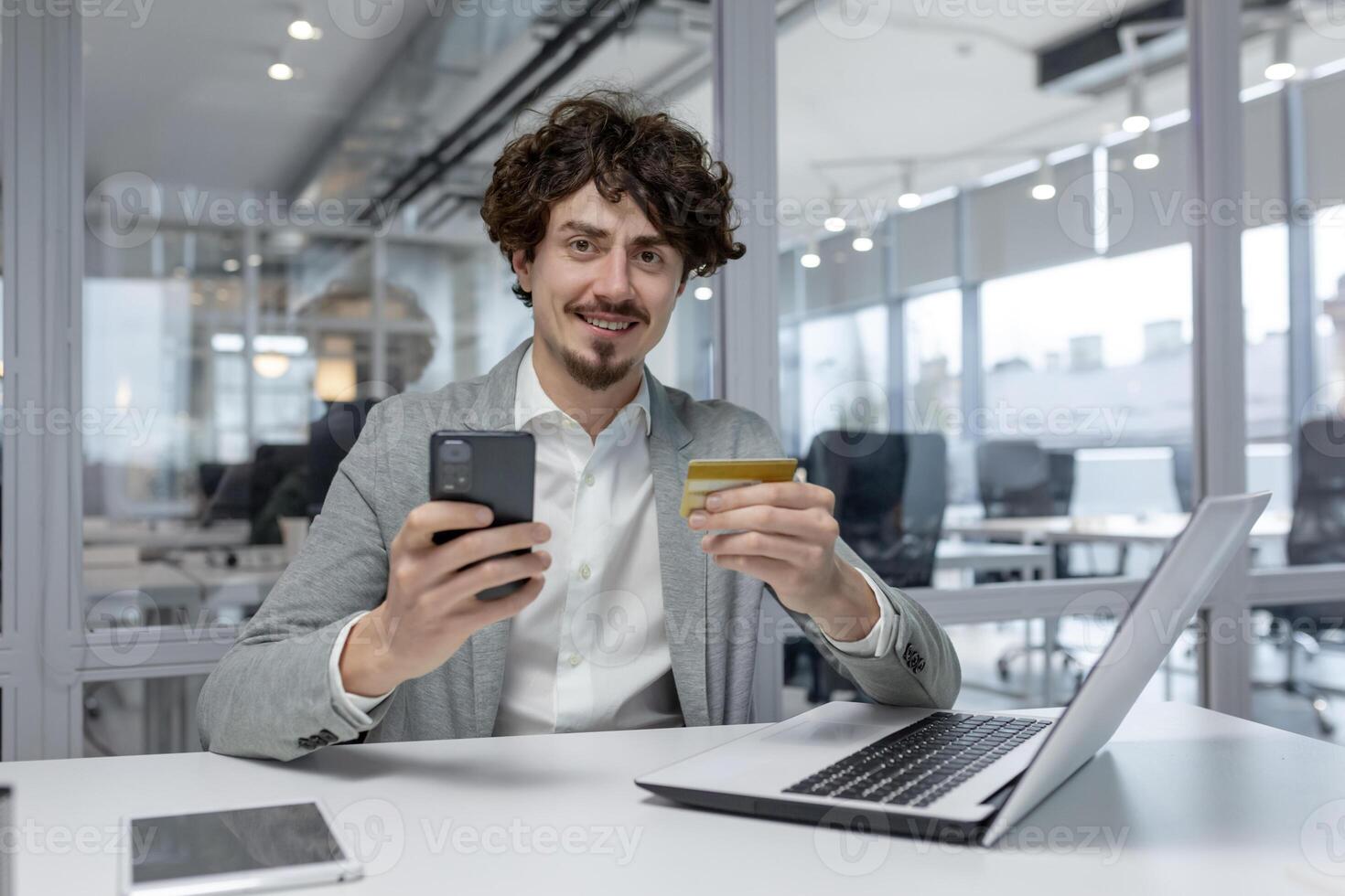 dai capelli ricci giovane uomo d'affari nel un' moderno ufficio multitasking con un' il computer portatile e smartphone, esemplificativo focalizzata aziendale opera. foto