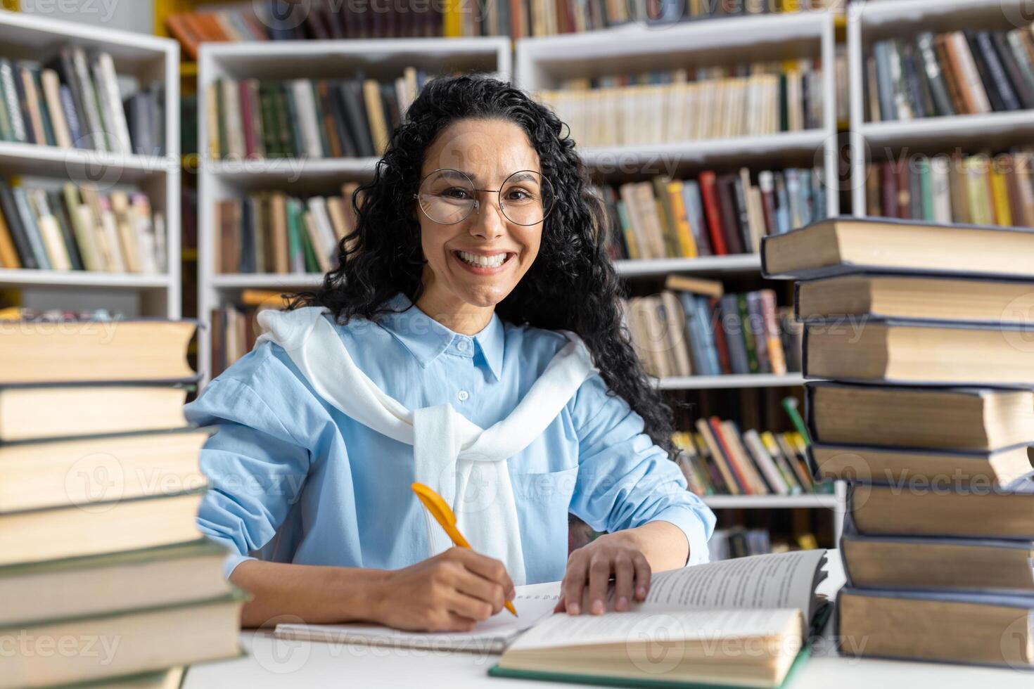 allegro ispanico femmina alunno con bicchieri studiando diligentemente tra torreggiante pile di libri nel un' biblioteca. foto