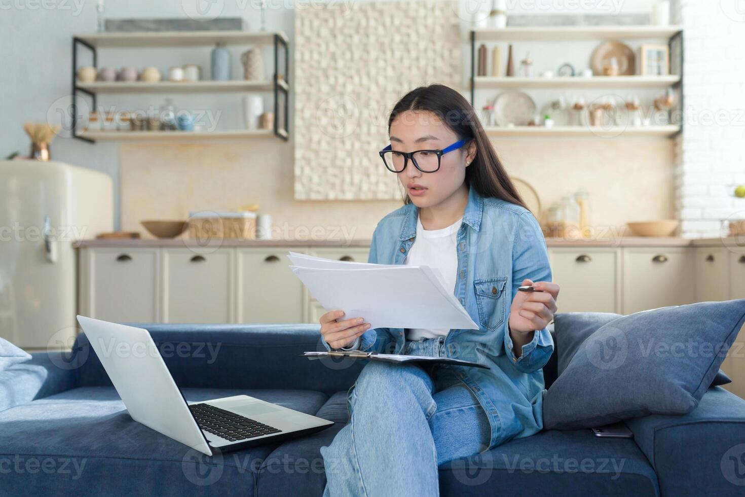 opera a casa in linea. occupato giovane asiatico donna architetto, progettista lavori a casa. lei si siede focalizzata su il divano con un' computer portatile, progetti, e un' penna. foto