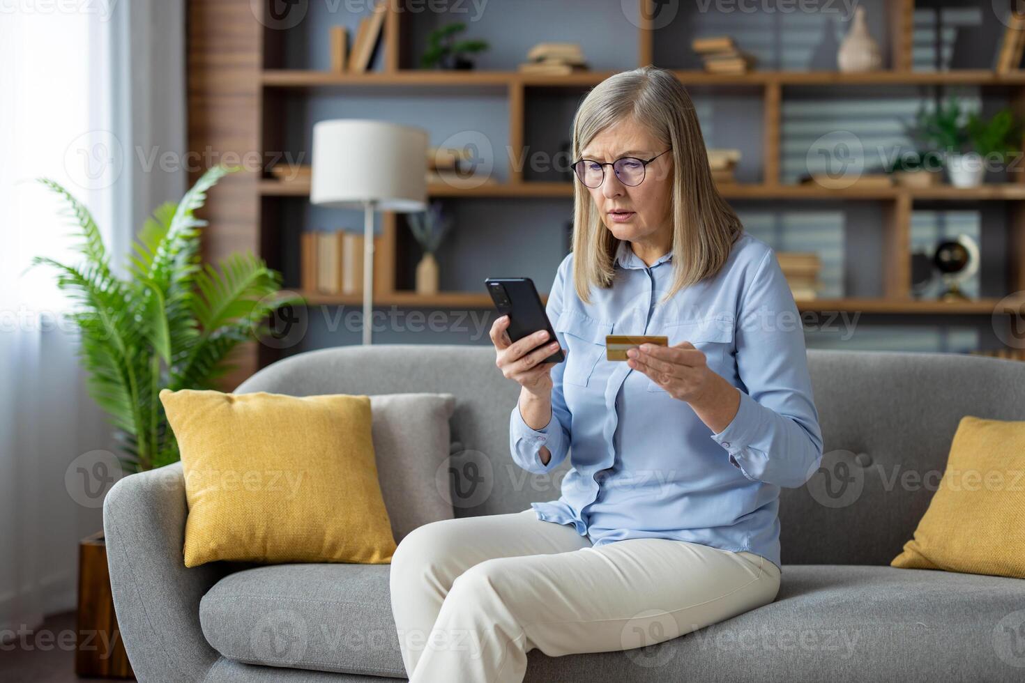 ansioso maturo donna seduta su divano, Tenere credito carta e Telefono, guardare a schermo con preoccupazione, sospettare un' fraudolento transazione. foto