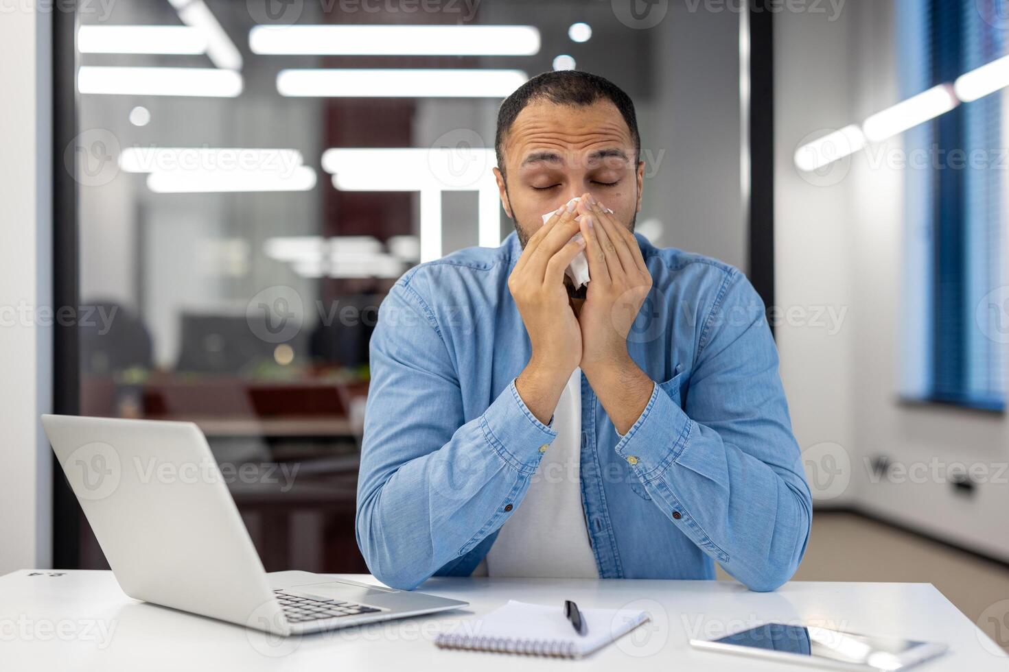 un' giovane latino americano uomo lavori nel il ufficio, si siede a un' scrivania e soffre a partire dal virale febbre e allergie, salviette il suo naso con un' tovagliolo a partire dal un' che cola naso. foto