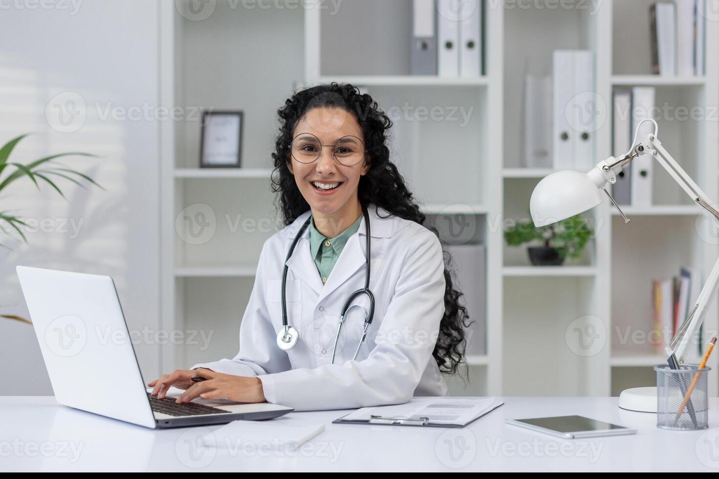 un' allegro latino femmina medico nel un' bianca cappotto sorrisi come lei lavori su sua il computer portatile nel un' moderno ufficio ambientazione. foto