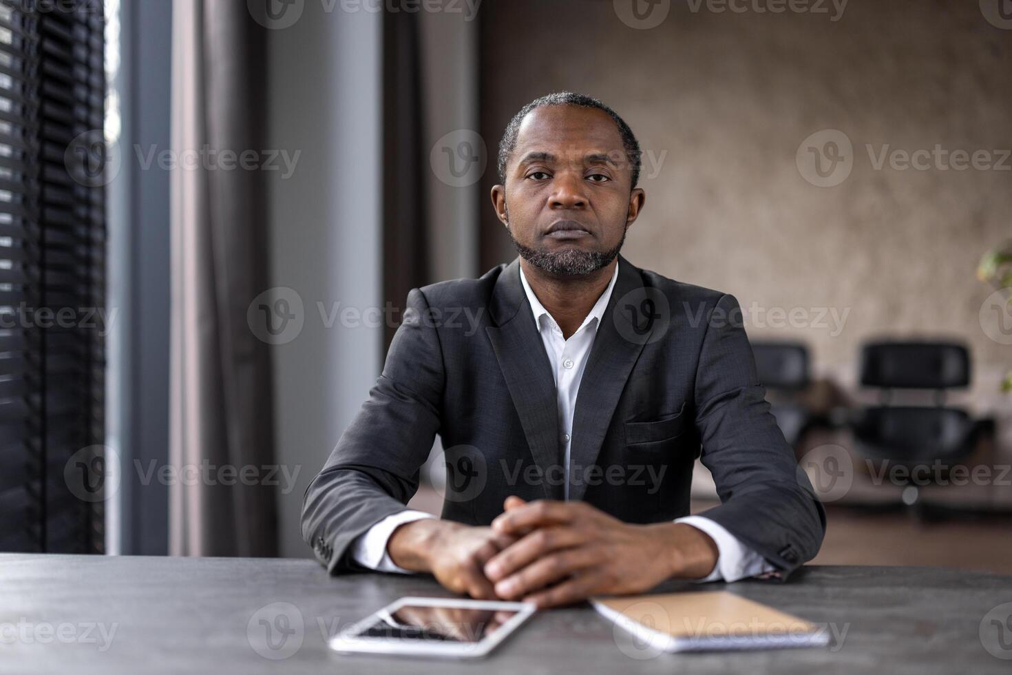 un' fiducioso africano americano uomo d'affari si siede a un' scrivania nel un' ben illuminato moderno ufficio, guardare direttamente a il telecamera, trasudante professionalità e messa a fuoco. foto