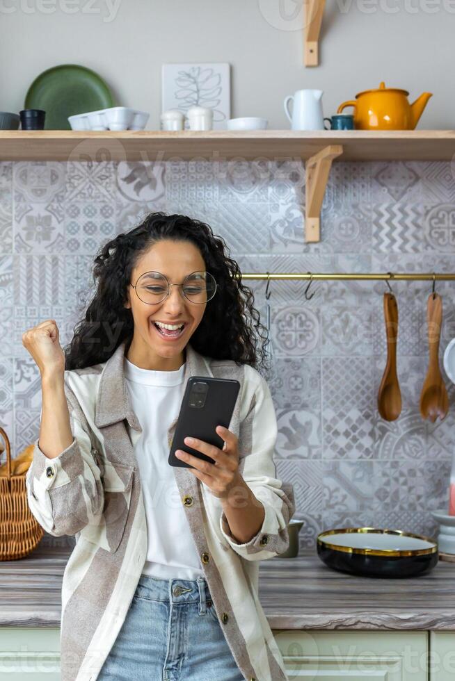 verticale sparo, latino americano donna a casa nel cucina avuto bene notizia in linea, donna con Riccio capelli e bicchieri festeggiare vittoria successo Tenere inteligente Telefono. foto