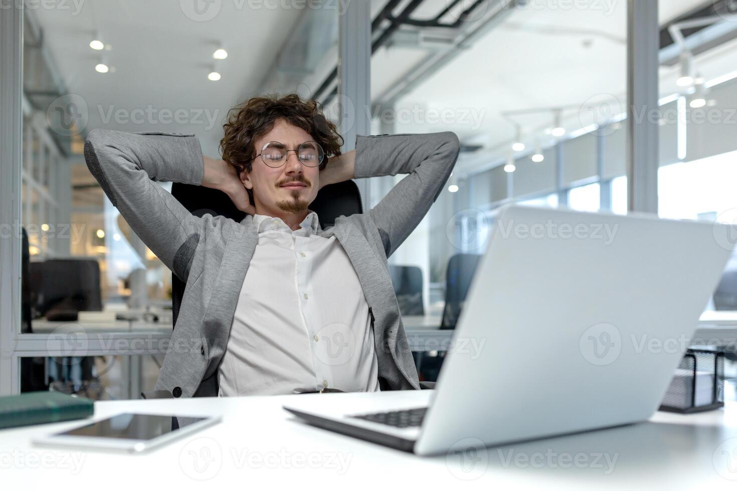 un' giovane uomo d'affari con Riccio capelli si distende nel un' moderno ufficio, mostrando un' momento di rilassamento in mezzo un' occupato aziendale ambientazione. foto