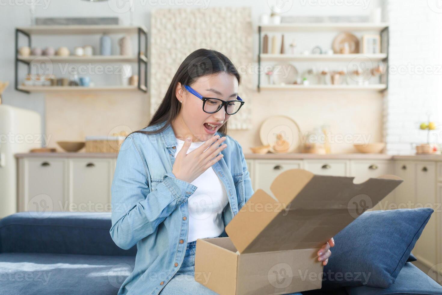 contento e piacevolmente sorpreso giovane bellissimo asiatico donna seduta su divano a casa e apertura cartone scatola, regalo. io ricevuto un inaspettato pacchetto nel il posta. foto