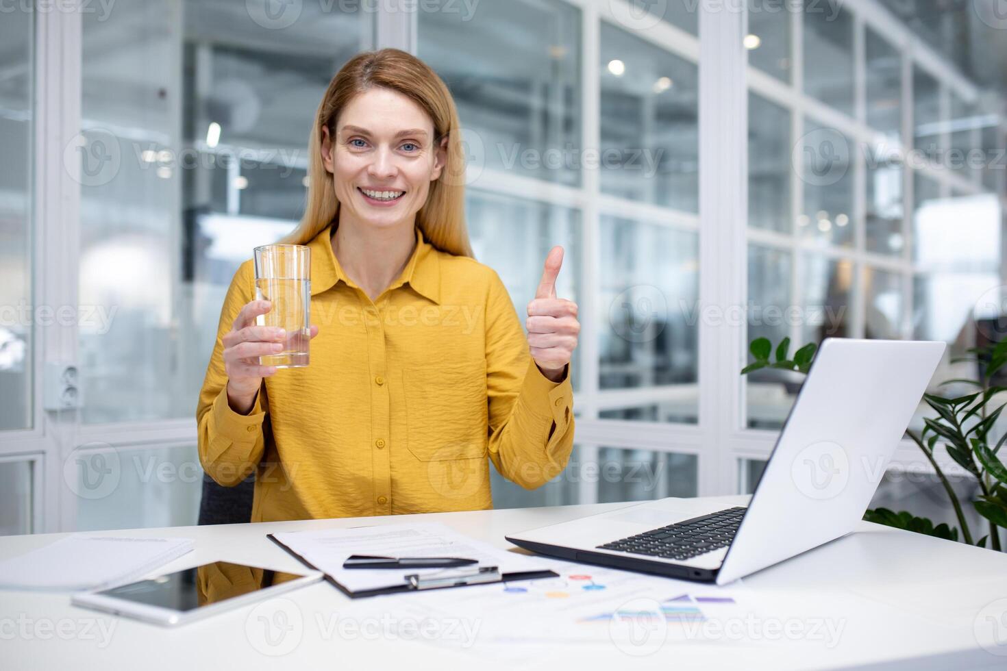 ritratto di un' giovane sorridente attività commerciale donna seduta a il scrivania nel il ufficio, Tenere un' bicchiere di acqua e puntamento super dito a il telecamera. foto