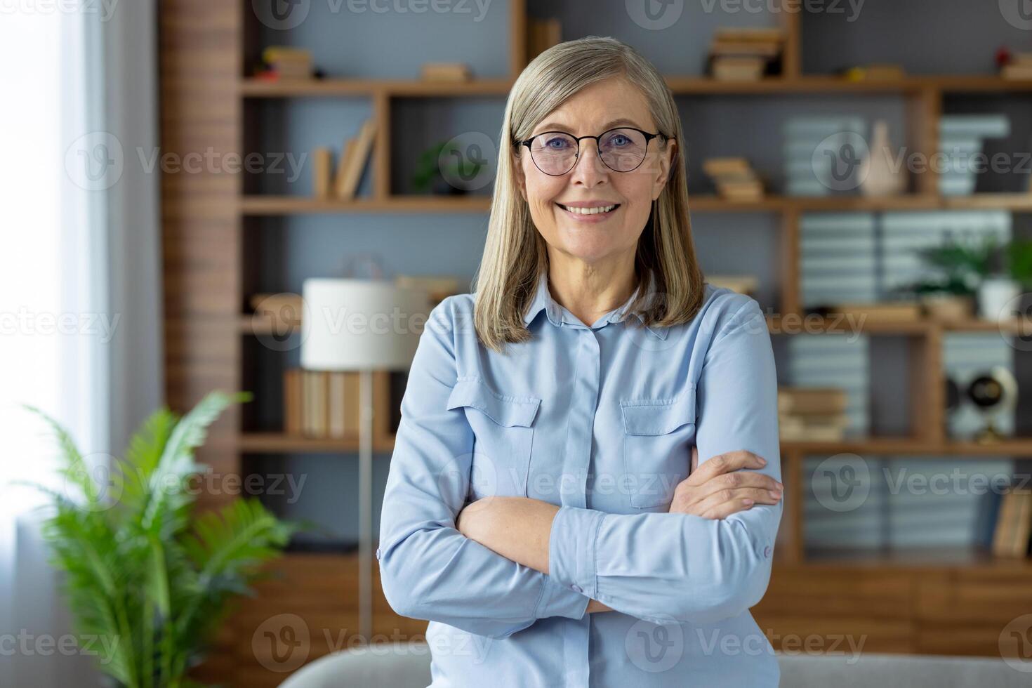 fiducioso femmina pensionato nel pastello camicetta in piedi con braccia attraversato su il petto e sorridente a telecamera. maturo signora nel spettacoli in posa a elegante appartamento interno e godendo vacanza al chiuso. foto