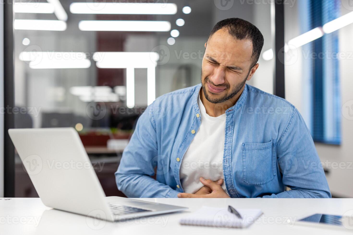 indiano uomo d'affari sperimentare stomaco dolore mentre Lavorando su un' il computer portatile nel un' moderno ufficio ambientazione, apparendo angosciato. foto