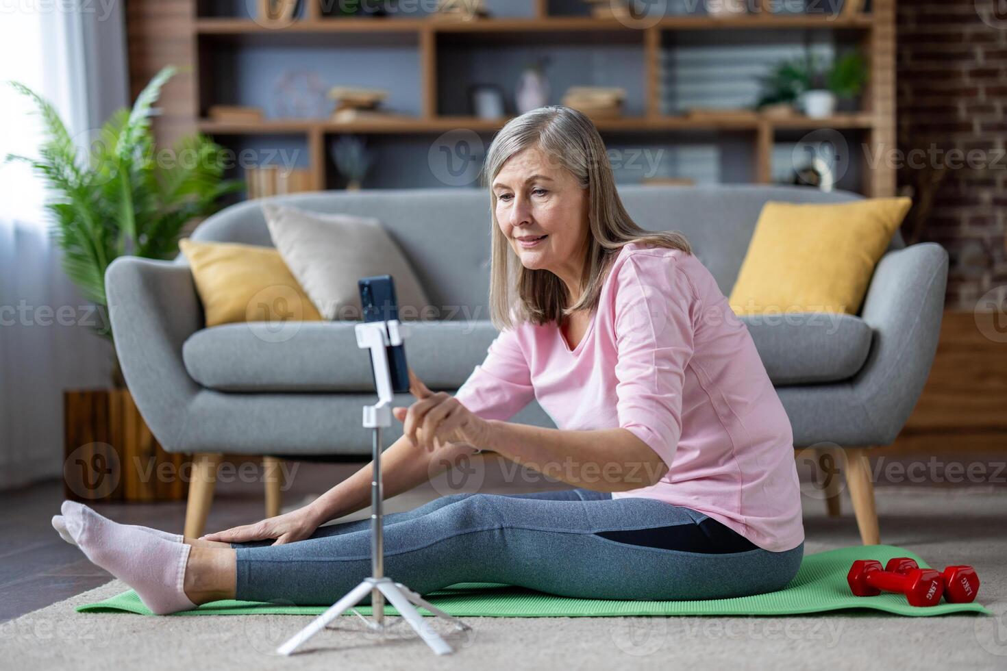 anziano maturo grigio dai capelli donna fare fitness seduta su esercizio stuoia a casa nel vivente camera su pavimento, utilizzando tripode tripode con Telefono, Guardando in linea classe e formazione . foto