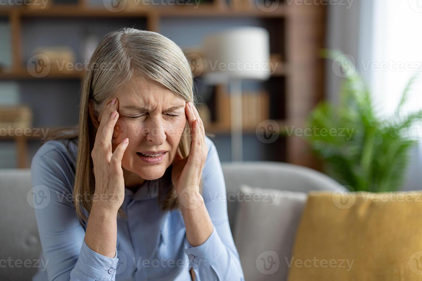 un' maturo donna si siede nel sua vivente camera, visibilmente afflitto con un' acuto male alla testa, Tenere sua templi nel dolore e disagio. foto