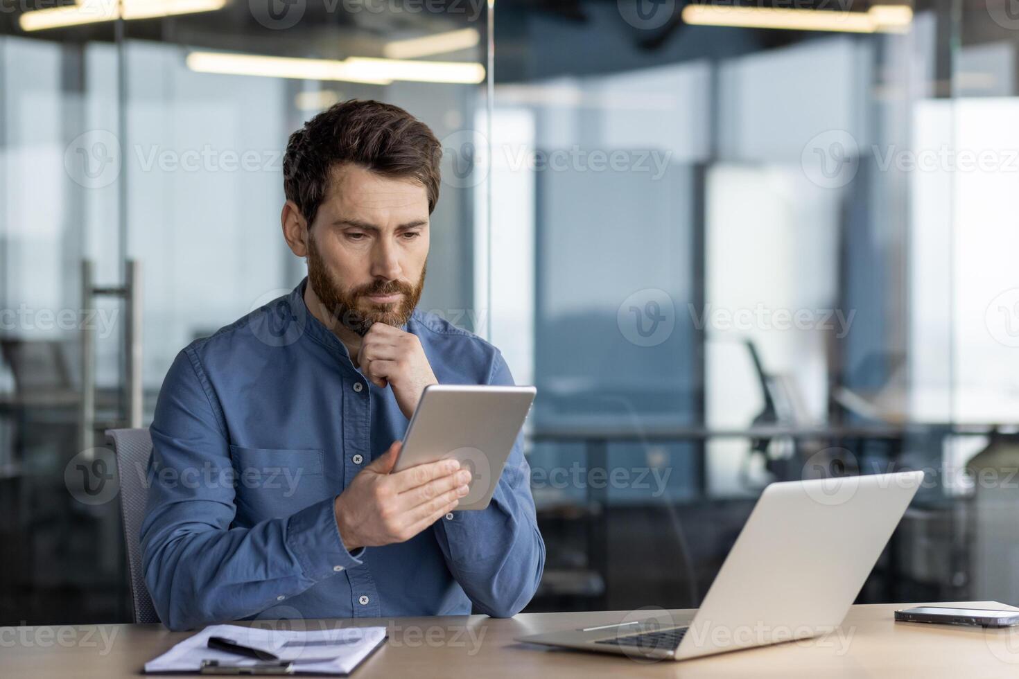 grave maschio professionale Lavorando su un' digitale tavoletta con un' il computer portatile e taccuino di il suo lato nel un' contemporaneo ufficio spazio. foto