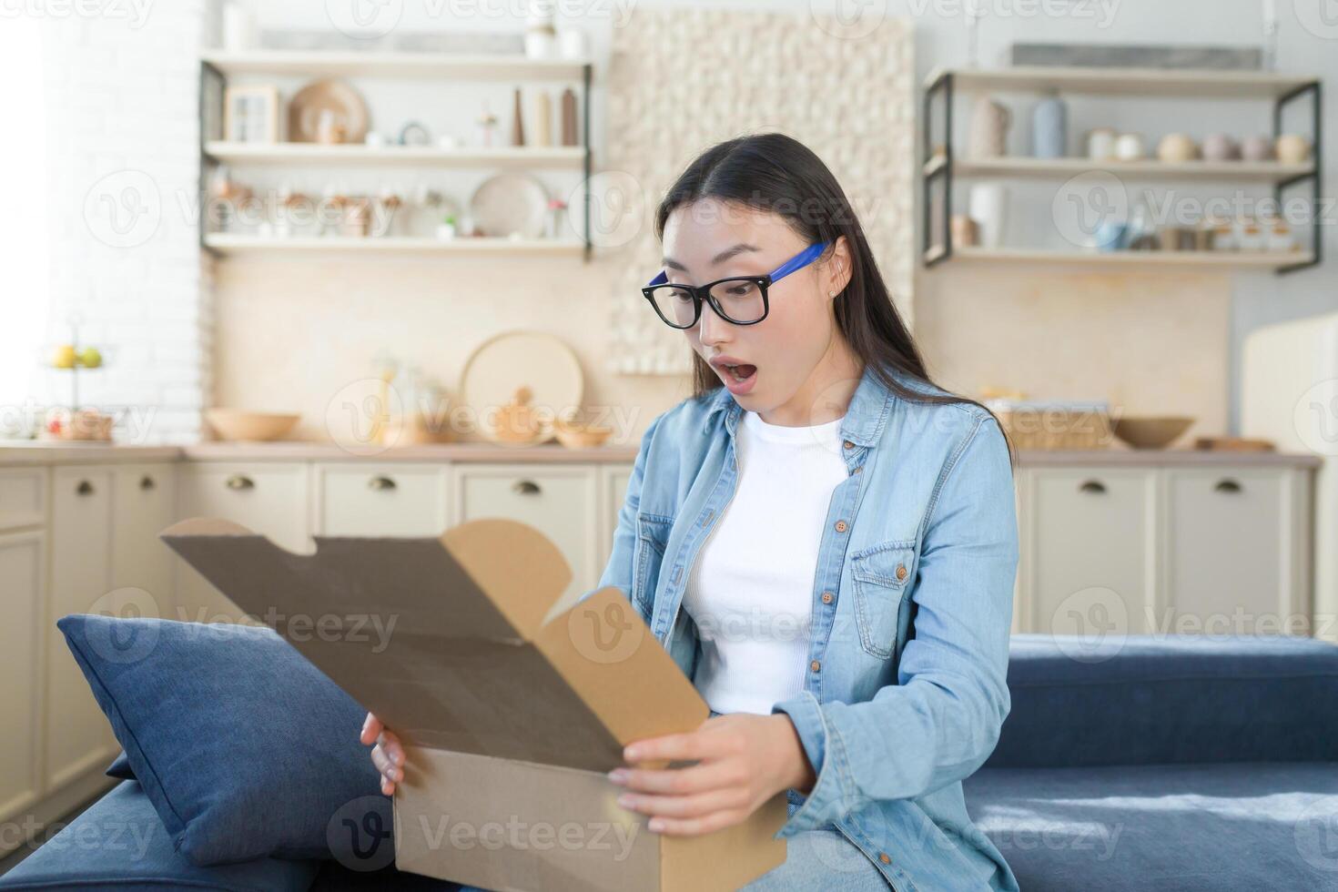 scioccato e allegro donna a casa ricevuto un' gioioso Acquista di un' regalo, si apre il scatola e esamina il regalo con soddisfazione, seduta su il cucina divano foto