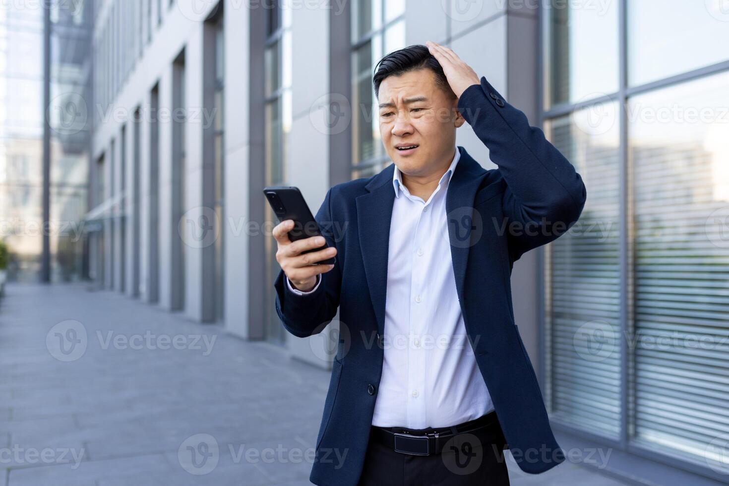 un' giovane asiatico uomo nel un' attività commerciale completo da uomo è in piedi al di fuori un ufficio centro, guardare scioccato e preoccupato a il Telefono schermo, e Tenere il suo testa. foto