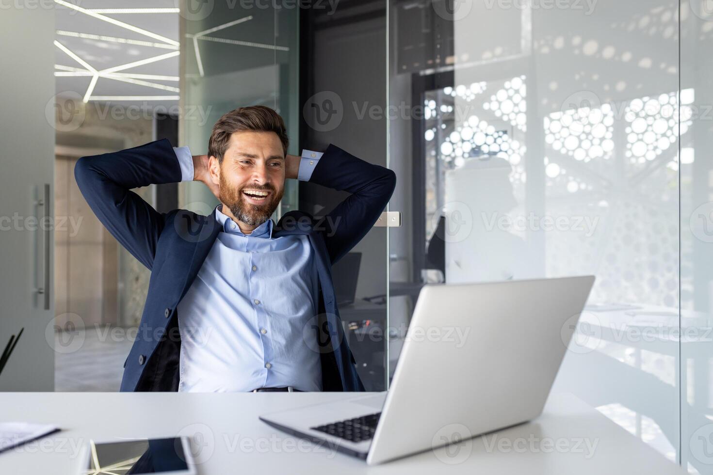 rilassato uomo d'affari con mani dietro a testa sorridente nel moderno ufficio, godendo successo e comfort, aziendale benessere. foto