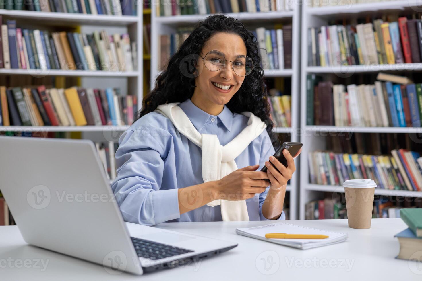 ritratto di un' bellissimo ispanico femmina alunno nel un' Università biblioteca tra libri, un' donna con Riccio capelli è sorridente e guardare a telecamera, Tenere affinare, utilizzando un applicazione in linea apprendimento. foto