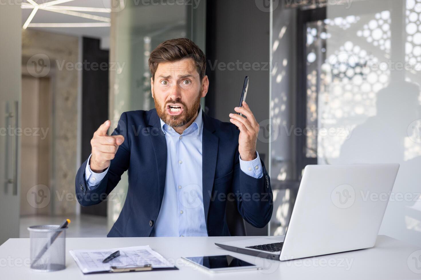 ritratto di un arrabbiato giovane uomo uomo d'affari seduta nel il ufficio a il tavolo, parlando su il Telefono, urlando e puntamento il suo dito a il telecamera accusando qualcuno. foto