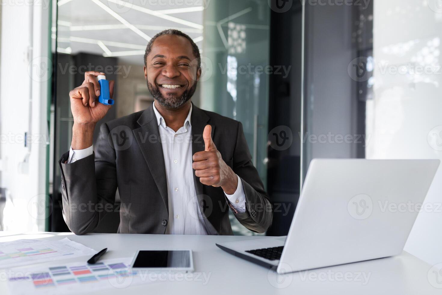 soddisfatto paziente dopo trattamento con asma inalatore, uomo dentro ufficio a posto di lavoro sorridente e guardare a telecamera, mostrando dito su e Tenere inalatore nel mani, foto