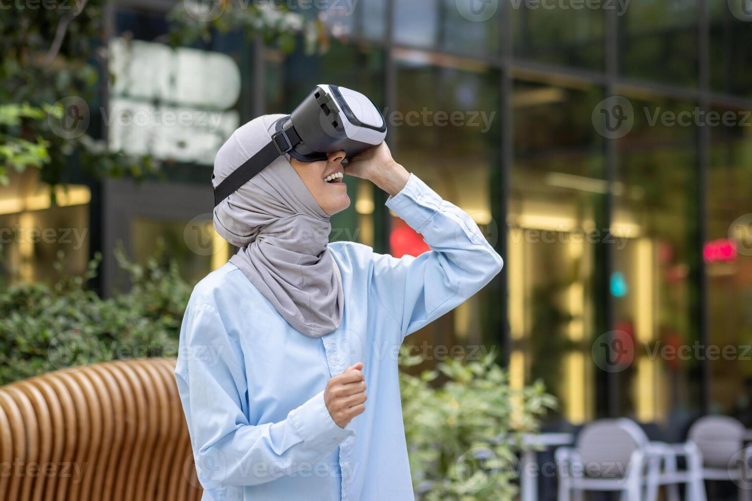 un' gioioso musulmano donna con un' hijab utilizzando un' virtuale la realtà cuffia nel un urbano ambientazione, mostrando eccitazione e curiosità. foto