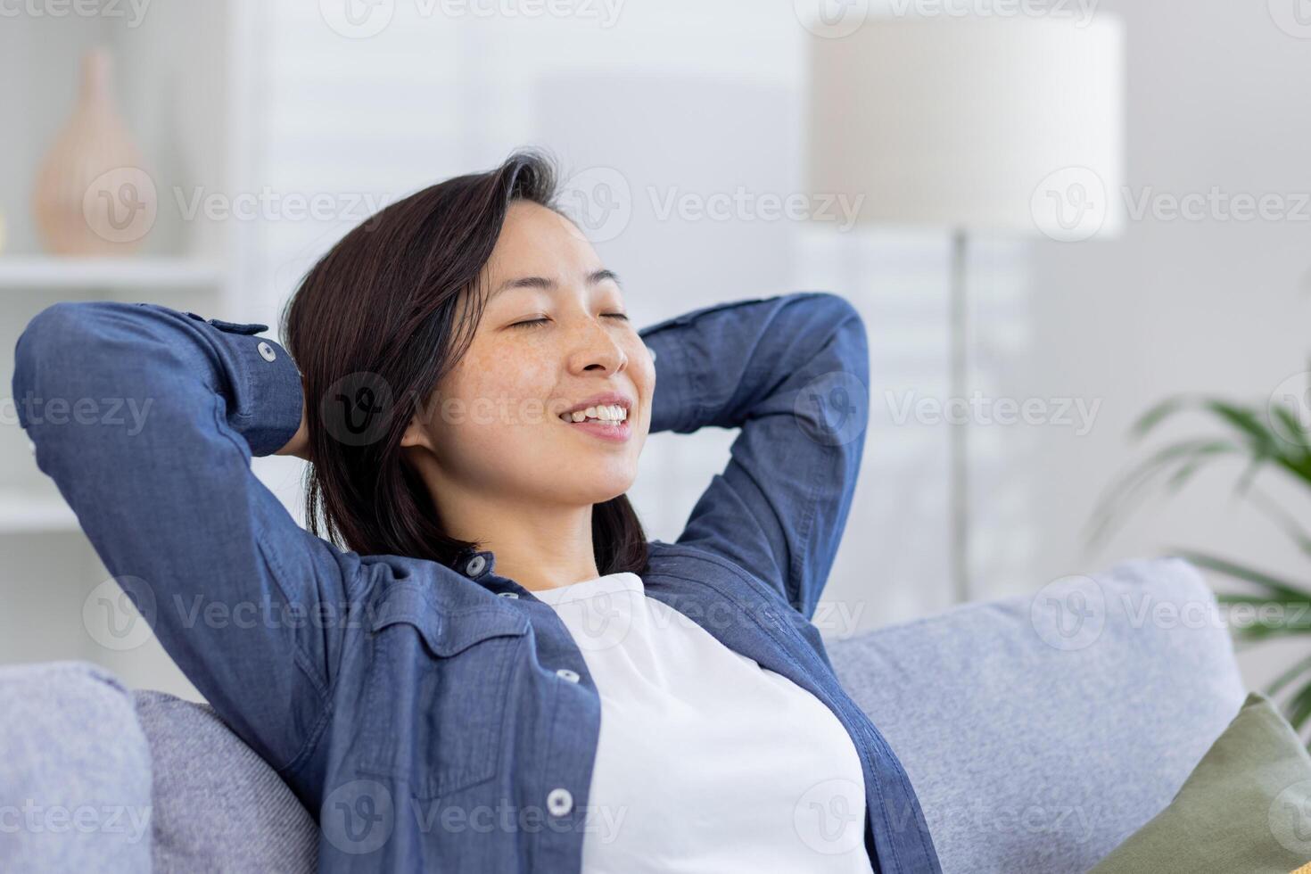 sognante asiatico donna rilassante a casa vicino su, donna sorridente e pensiero seduta su divano con mani dietro a testa con chiuso occhi. foto
