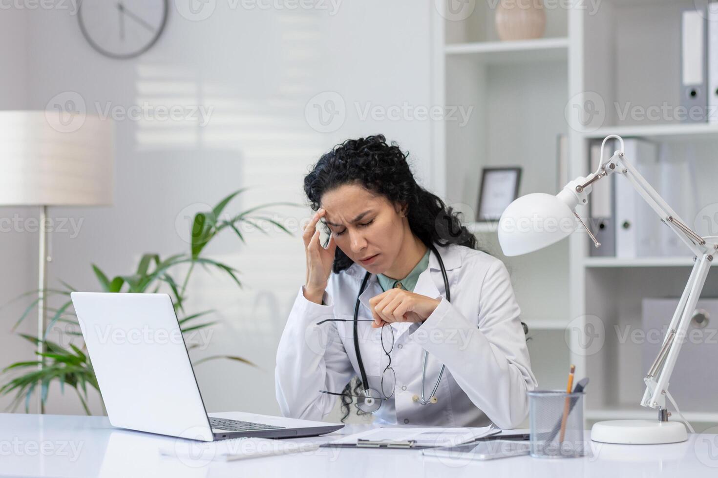 un' preoccupato ispanico femmina medico appare stressato mentre parlando su il Telefono nel sua ufficio, trasudante un aria di preoccupazione e concentrazione. foto