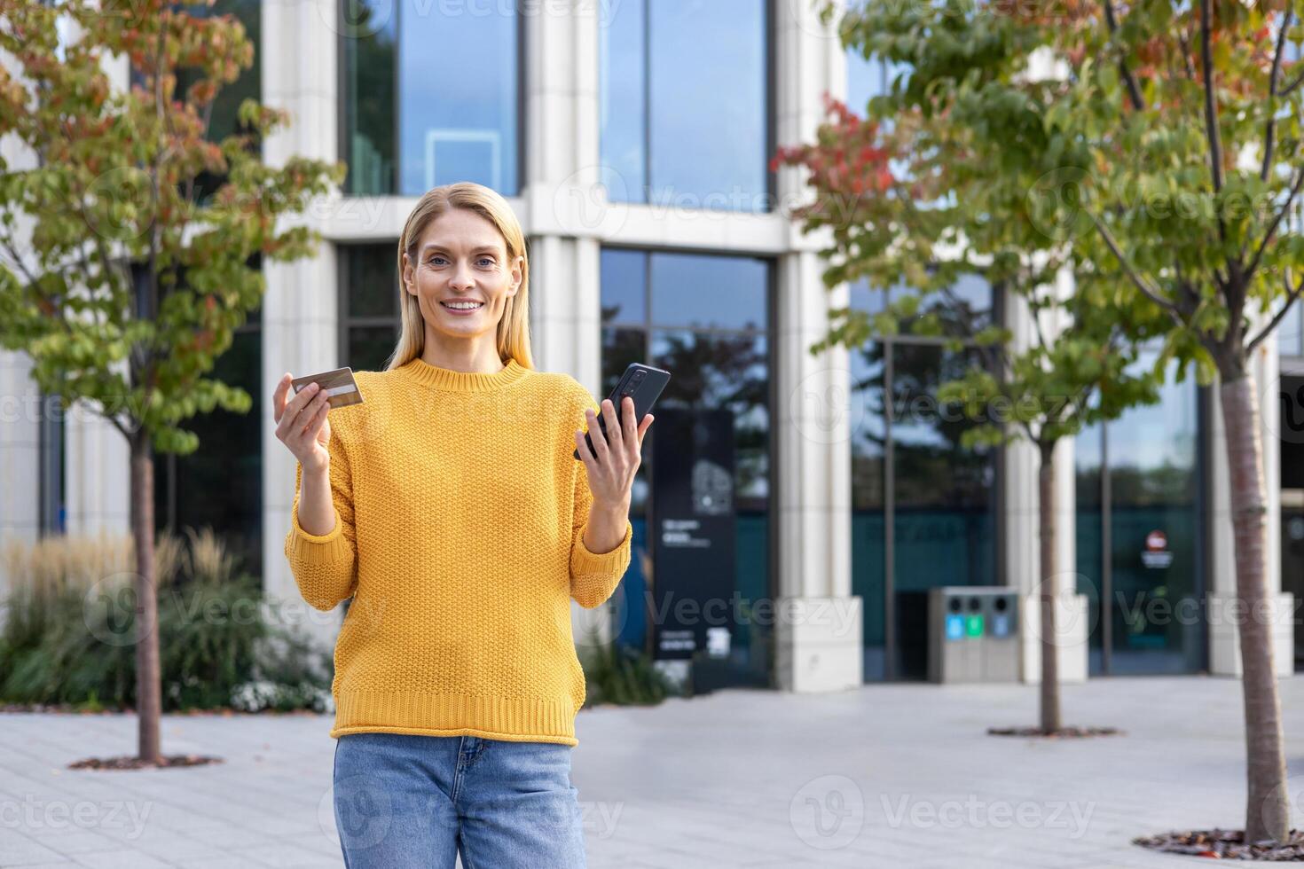 un' allegro bionda donna nel un' giallo maglione detiene un' credito carta e un' smartphone al di fuori un' moderno edificio con alberi nel il sfondo. foto