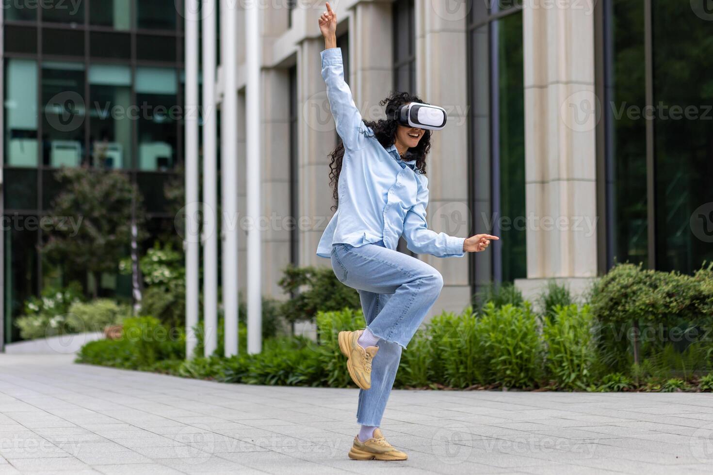 giovane bellissimo ragazza nel camicia e jeans danza al di fuori ufficio edificio indossare virtuale la realtà occhiali, ispanico donna contento avendo divertimento e rilassante con Riccio capelli. foto