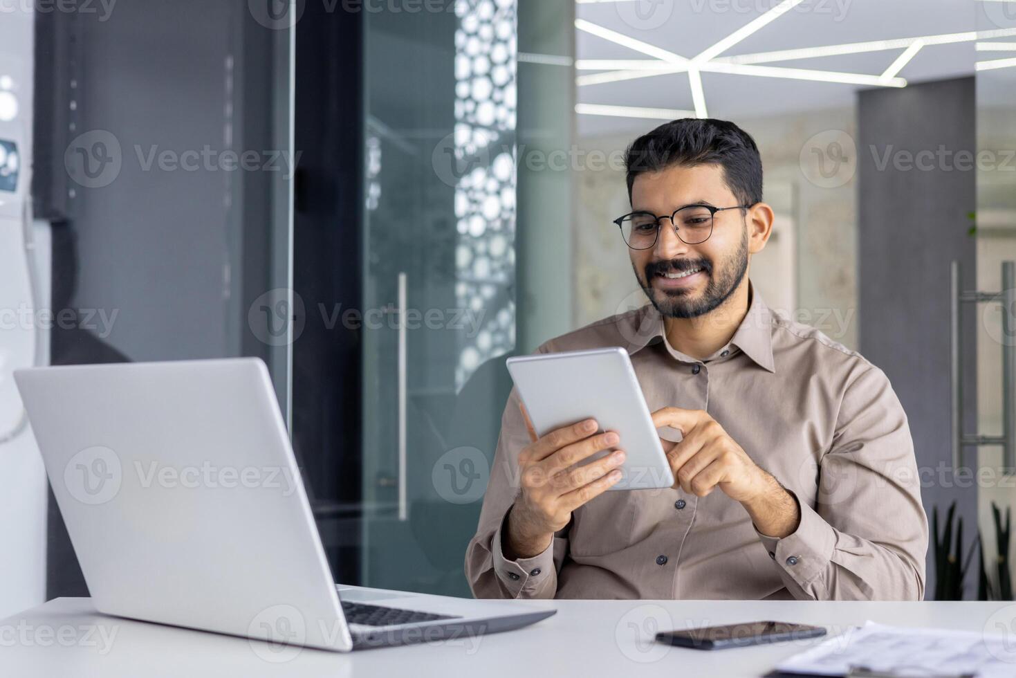 un' allegro indiano programmatore è sorridente e utilizzando un' digitale tavoletta, seduta a il suo scrivania nel un' moderno ufficio ambientazione. foto