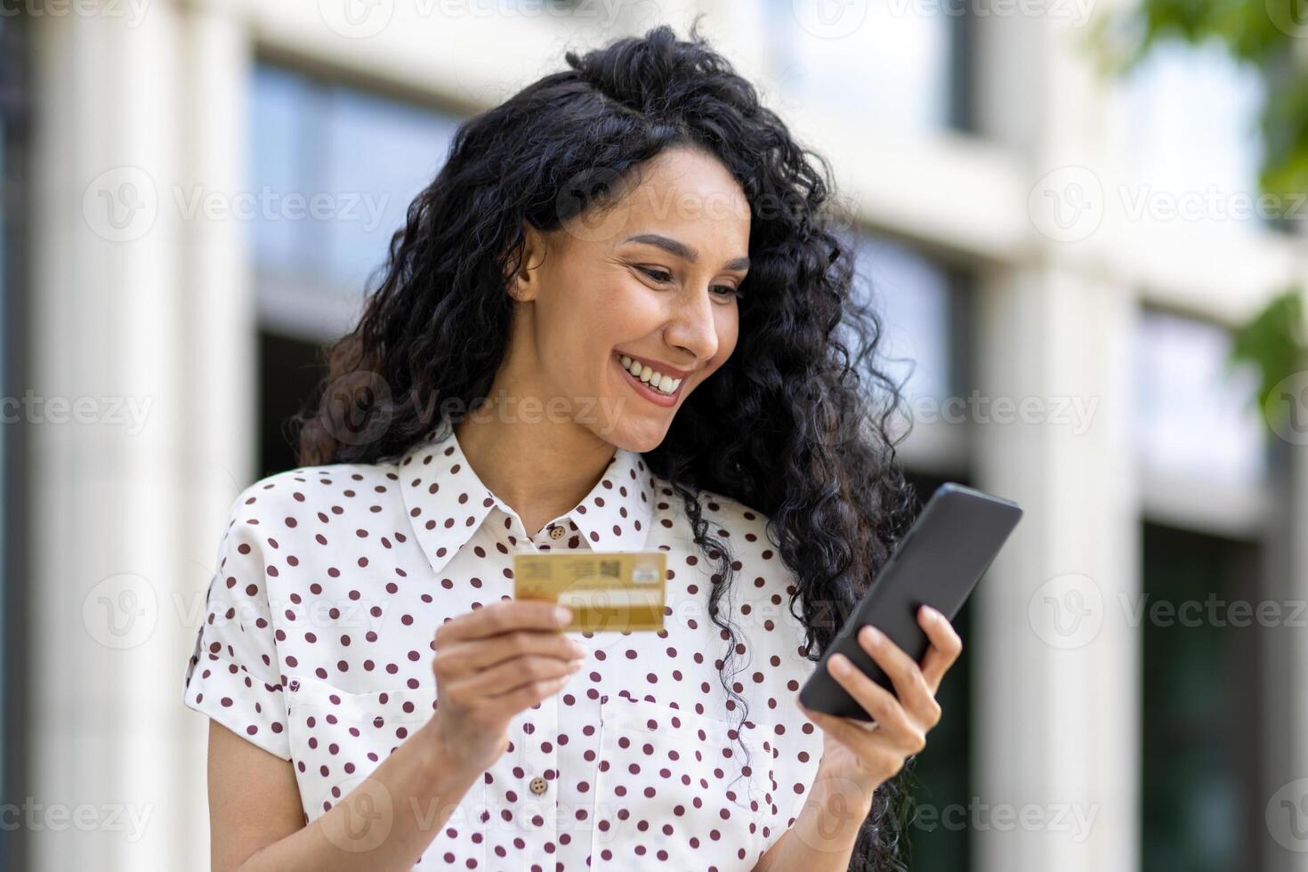 giovane bellissimo donna a piedi nel il città, soddisfatto latino americano donna con Riccio capelli detiene Telefono e banca credito carta nel mani, fa in linea acquisti felicemente libri Servizi e sceglie in linea. foto