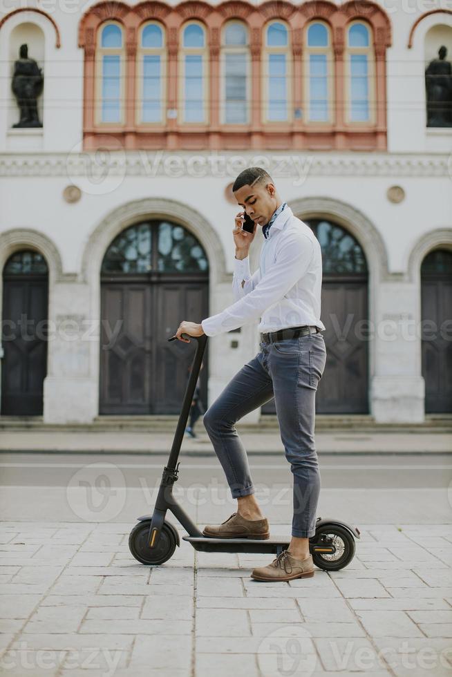 giovane afroamericano che utilizza il telefono cellulare mentre sta in piedi con uno scooter elettrico su una strada foto