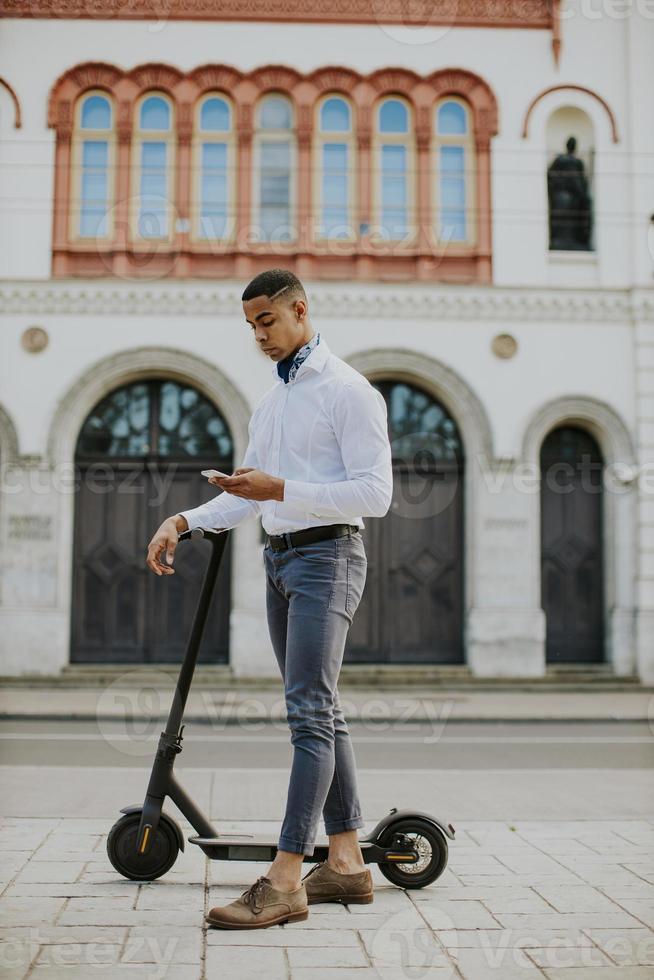 giovane afroamericano che utilizza il telefono cellulare mentre sta in piedi con uno scooter elettrico su una strada foto