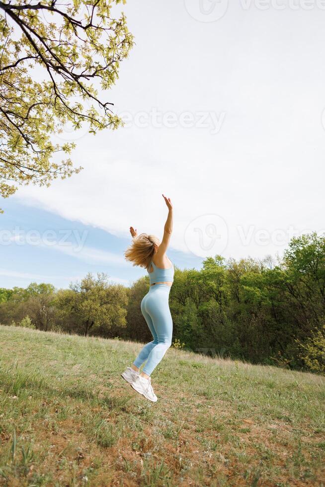 un atletico ragazza esegue il esercizio di salto su con sua braccia sollevato. bellissimo bionda caucasico donna nel blu stretto tuta. bionda ragazza a un all'aperto formazione sessione foto