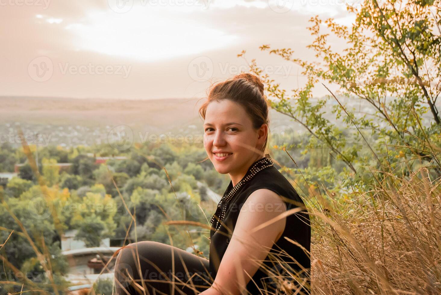 un' sorridente donna seduta nel natura a tramonto, godendo il tranquillo paesaggio foto