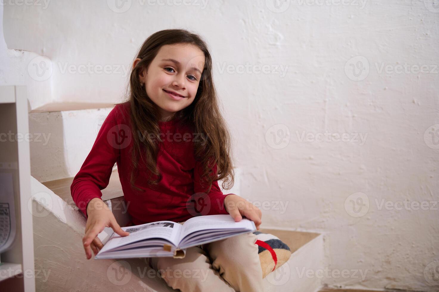 inteligente primario scuola alunno ragazza sorridente a telecamera, seduta su passaggi a casa con un' libro nel mani. poco ragazza lettura un' libro. mondo libro giorno concetto. indietro per scuola su nuovo semestre di accademico anno foto