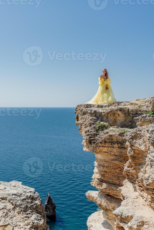 donna nel un' giallo vestito su il mare. lato Visualizza giovane bellissimo sensuale donna nel giallo lungo vestito in posa su un' roccia alto sopra il mare a tramonto. ragazza nel natura contro il blu cielo foto