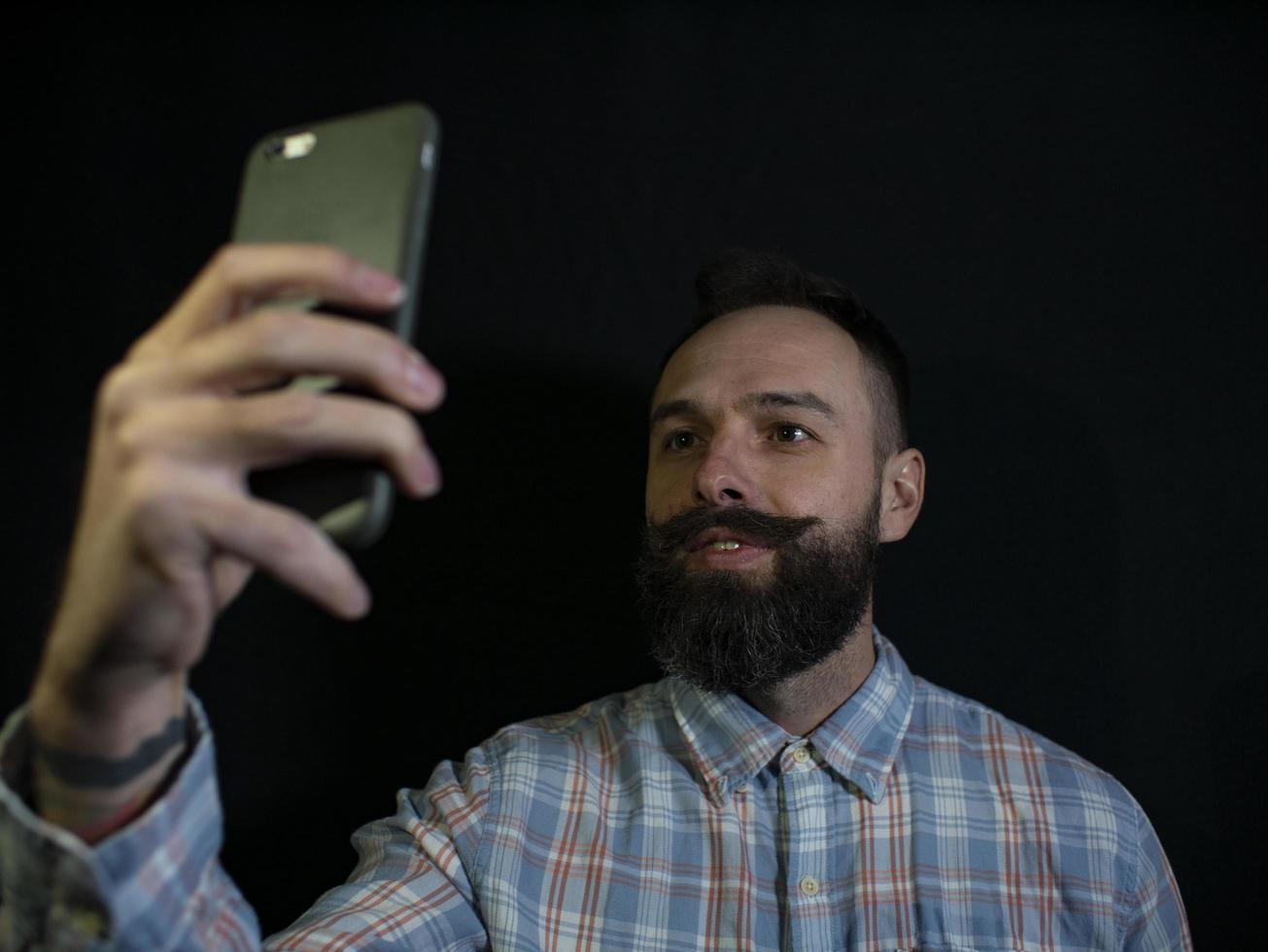 uomo elegante con barba e baffi fa selfie al telefono su sfondo nero foto