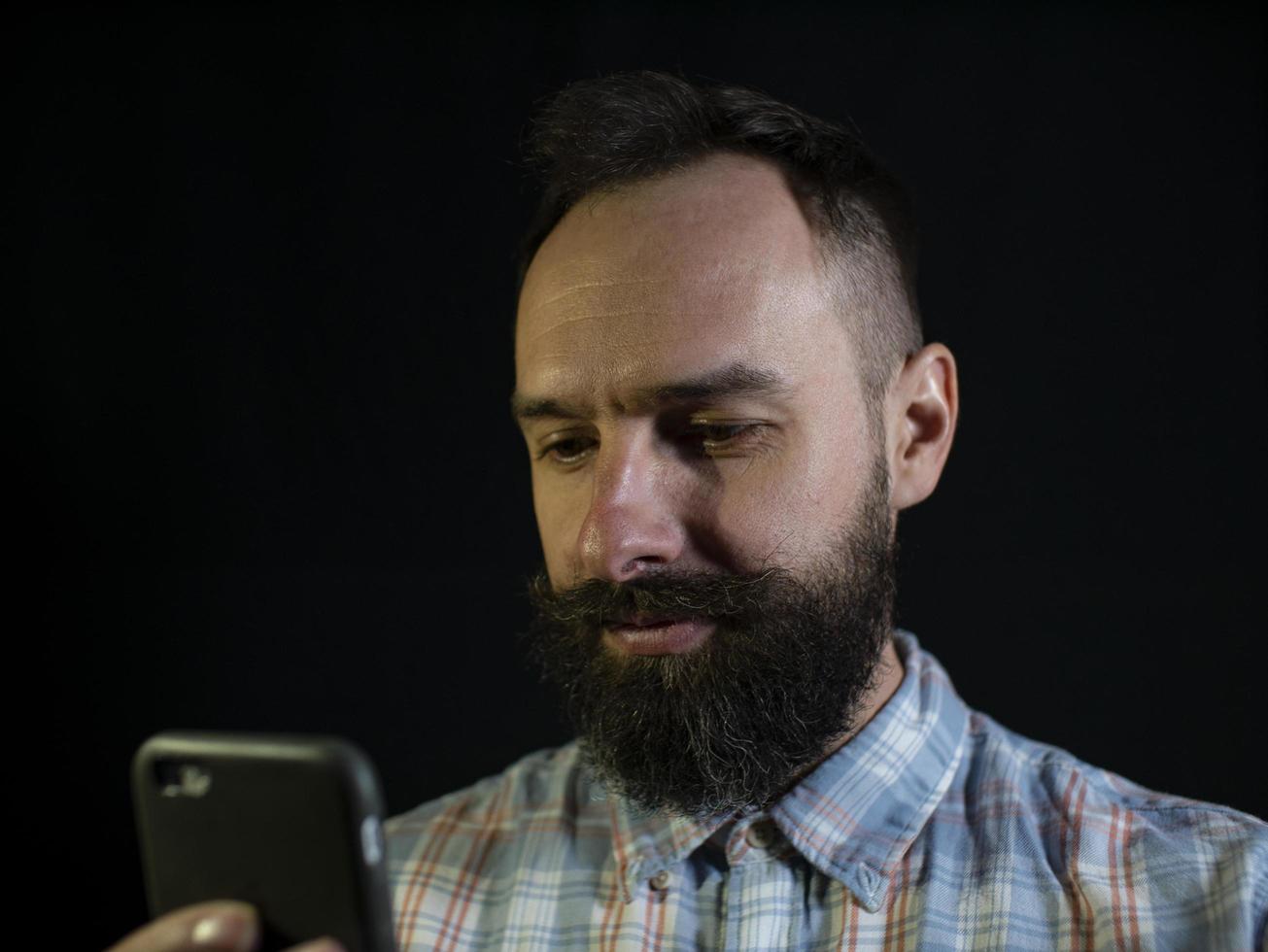 uomo elegante con barba e baffi guarda attentamente il telefono su sfondo nero foto